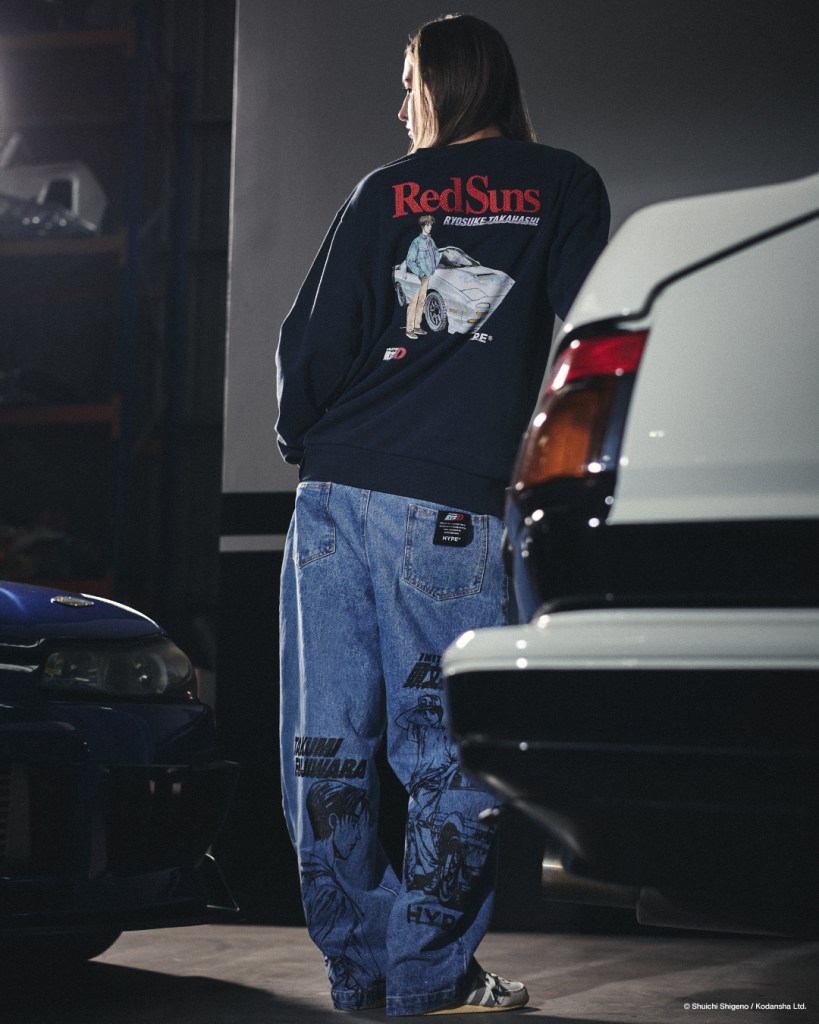 A person wearing a black sweatshirt featuring the 'RedSuns' design and blue baggy jeans with graphics, standing near classic cars in a dimly lit garage.
