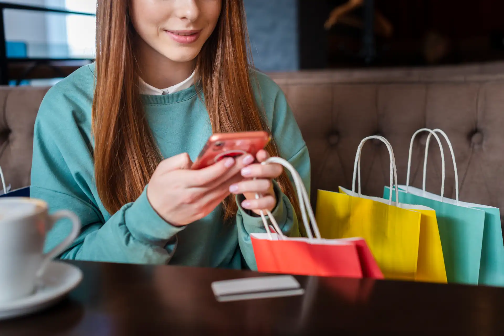 Woman with colorful shopping bags checking smartphone in a cafe, illustrating the trend of ambient shopping and mobile commerce.