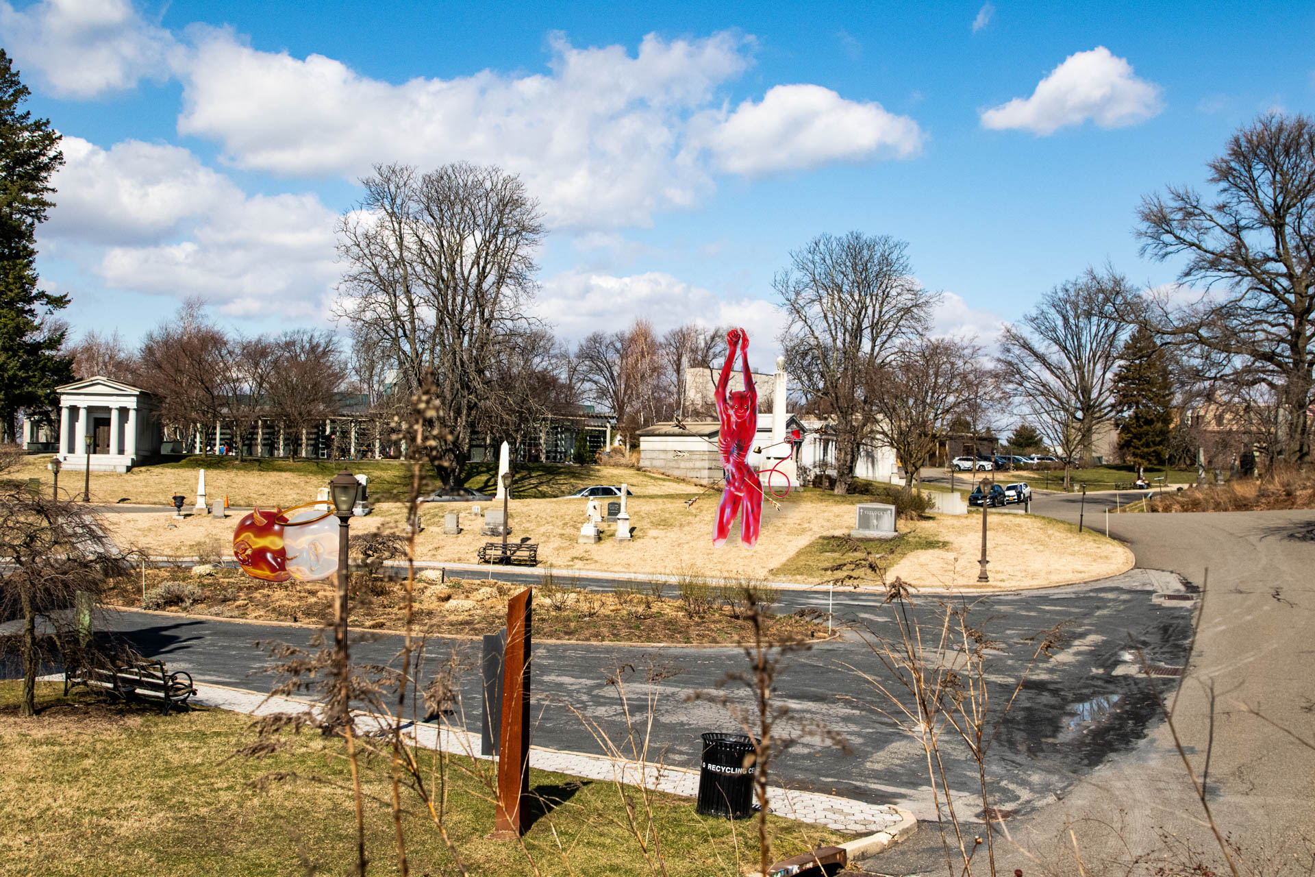 The image shows a park scene under a mostly clear blue sky with some white clouds. A large, red, abstract sculpture of a human figure dominates the center of the frame. The sculpture is surrounded by bare trees, giving the impression of a winter or early spring season. In the background, a white building with columns is visible, along with other structures including a section of a road or pathway. The ground in the park is mostly bare, with patches of dry grass and some bare earth. A dark-colored trash can or receptacle is visible near the bottom right. The photo is taken from a slightly elevated angle, offering a view of the park and its features.
