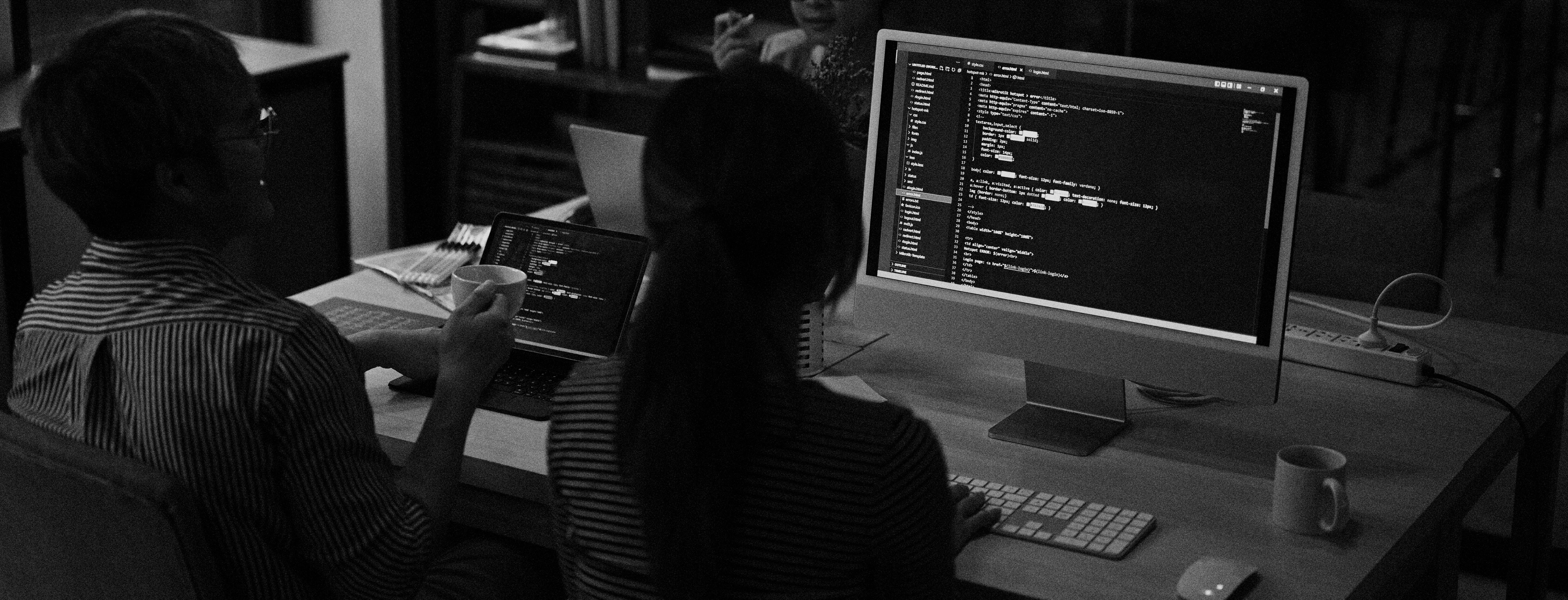 Una fotografía en blanco y negro de un espacio de trabajo colaborativo con tres personas sentadas alrededor de un escritorio. El enfoque está en dos computadoras que muestran entornos de codificación, una siendo una laptop y la otra un monitor de escritorio. La pantalla de escritorio muestra líneas de código HTML y CSS, mientras que la pantalla de la laptop exhibe código similar. El ambiente parece ser informal, con tazas de café y una disposición casual de teclados y periféricos, lo que sugiere un equipo trabajando en un proyecto de programación en un entorno relajado.