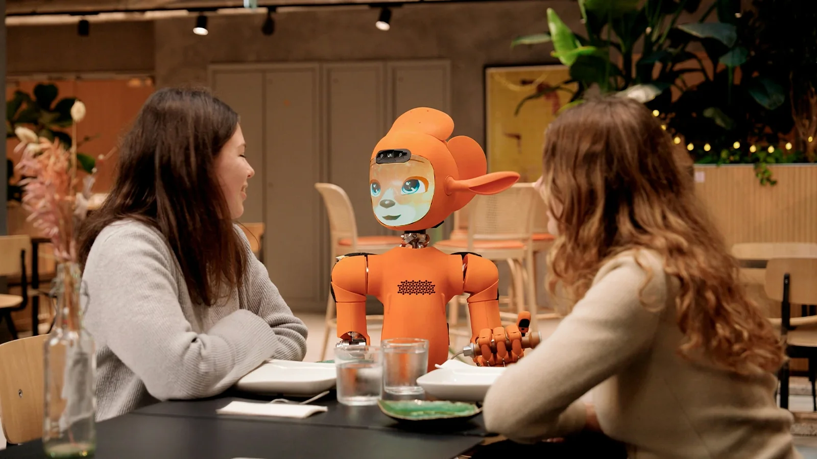 Orange robot with blue eyes sits at table between two women in modern restaurant setting.