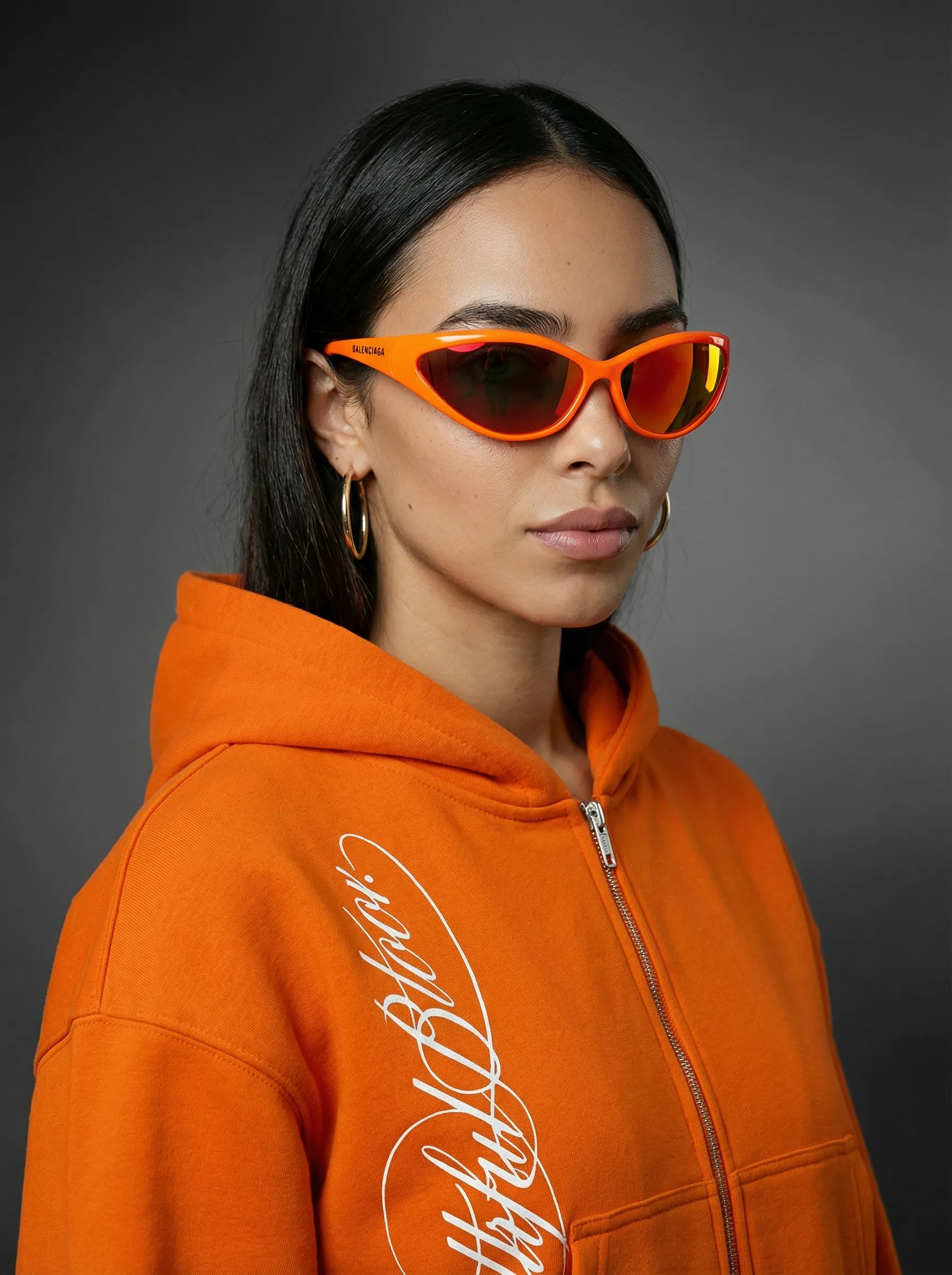 A person wearing vibrant orange sunglasses and a matching hoodie stands against a dark gray background, exuding a modern, stylish appearance.
