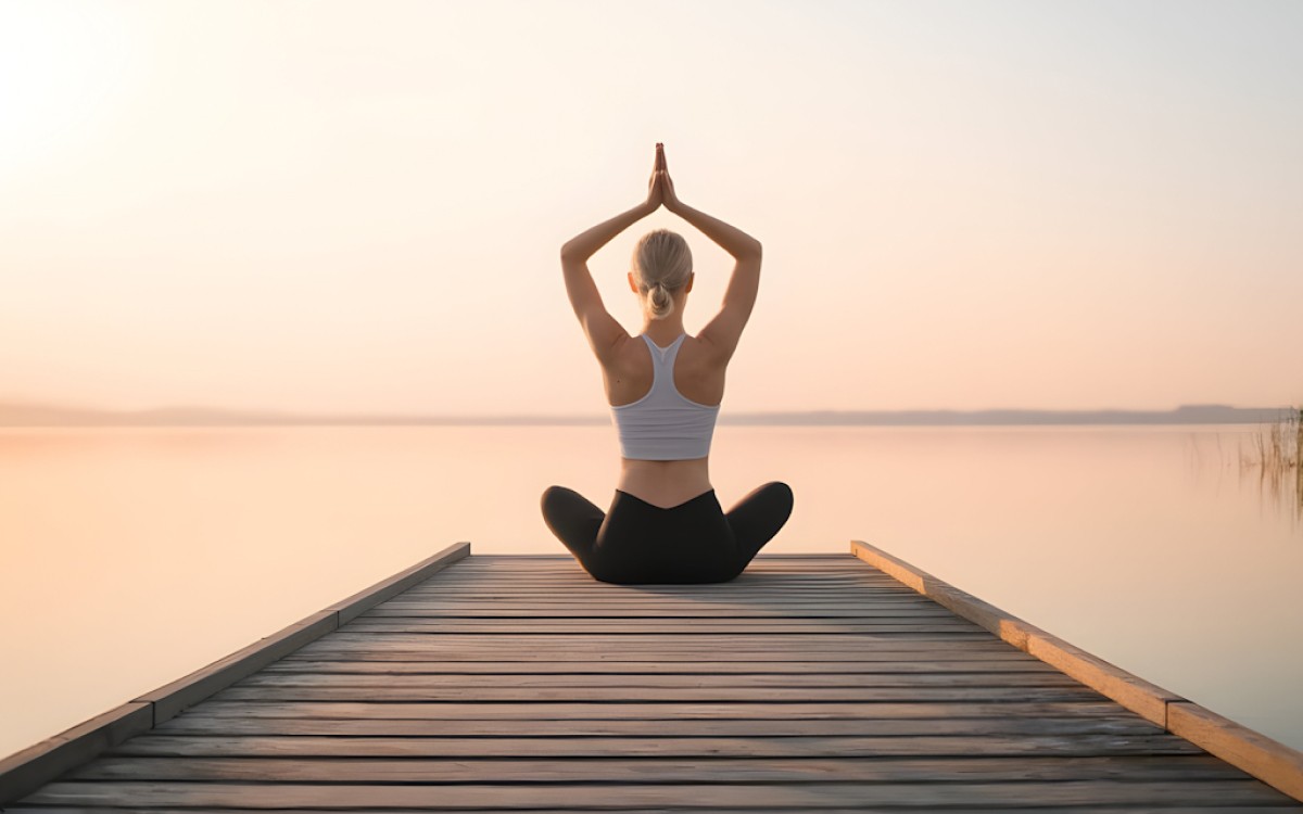 A woman practices yoga, sitting cross-legged on a wooden pier at sunrise, by a calm lake. She raises her hands above her head in a peaceful pose.