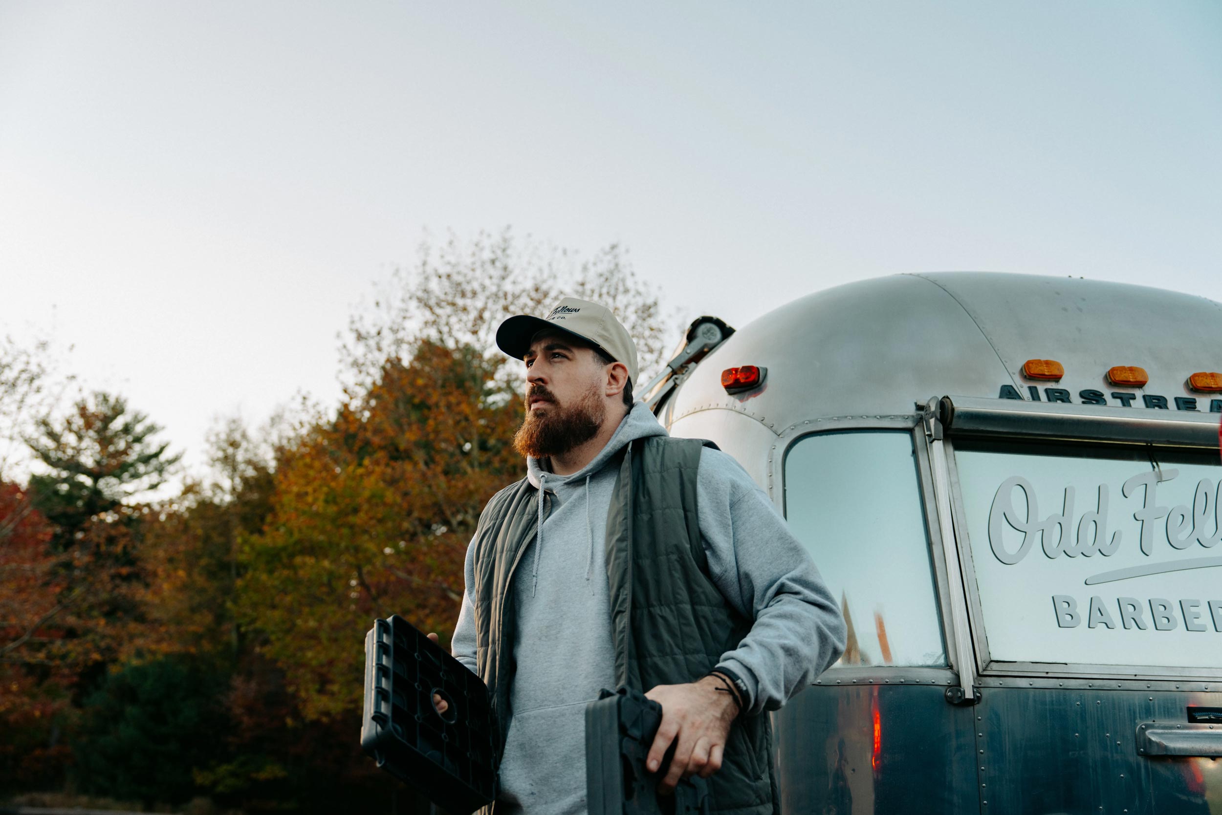 A bearded man in a baseball cap, grey hoodie, and dark vest stands outdoors, looking up and away from the camera, next to a silver Airstream-style trailer. The trailer has the words "Odd Fellows BARBER" on its side.