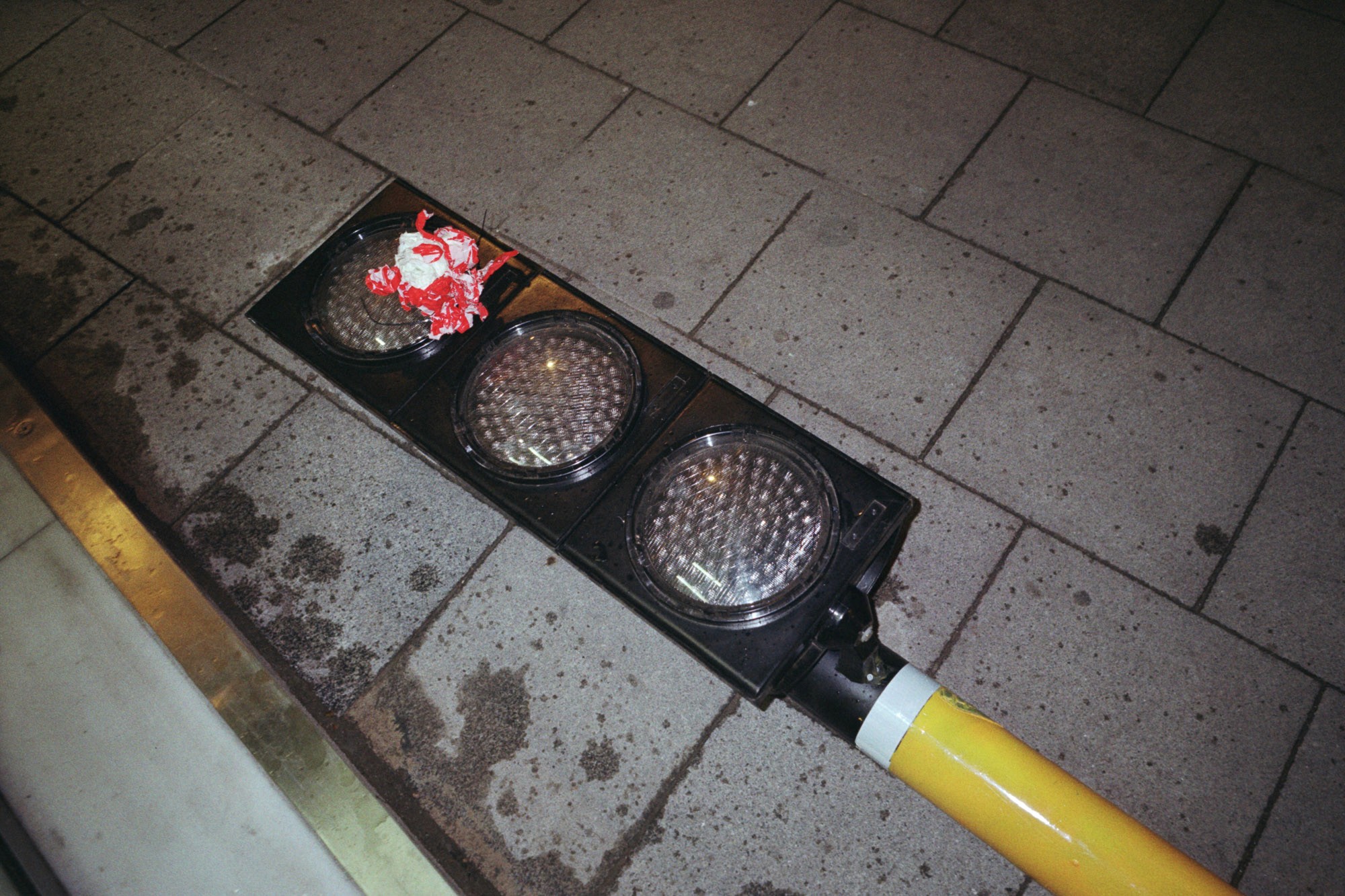 A fallen traffic light lies on the sidewalk, with a smashed red lens and a yellow pole, illustrating street maintenance issues.