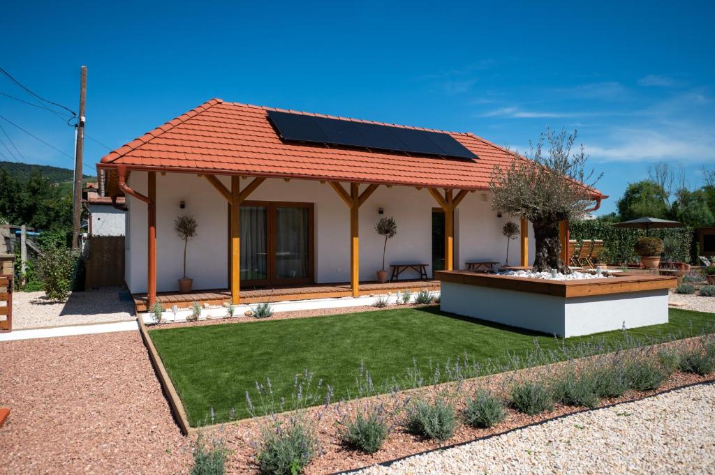 Egy modern, egyszintes fehér ház piros nyeregtetővel és napelemekkel, gondozott gyeppel, kavicsos sétányokkal és az előtérben magasított kerti ágyással, ragyogó kék ég alatt.