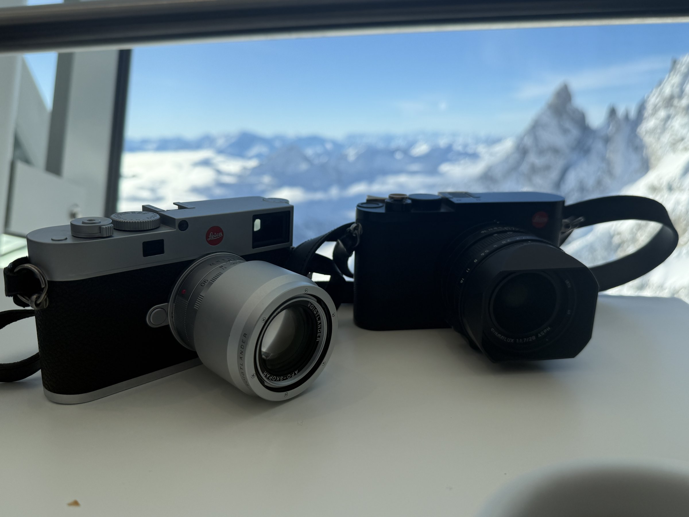 Two Leica cameras on a table before a window overlooking snowy Mont Blanc peaks near Courmayeur, Italy