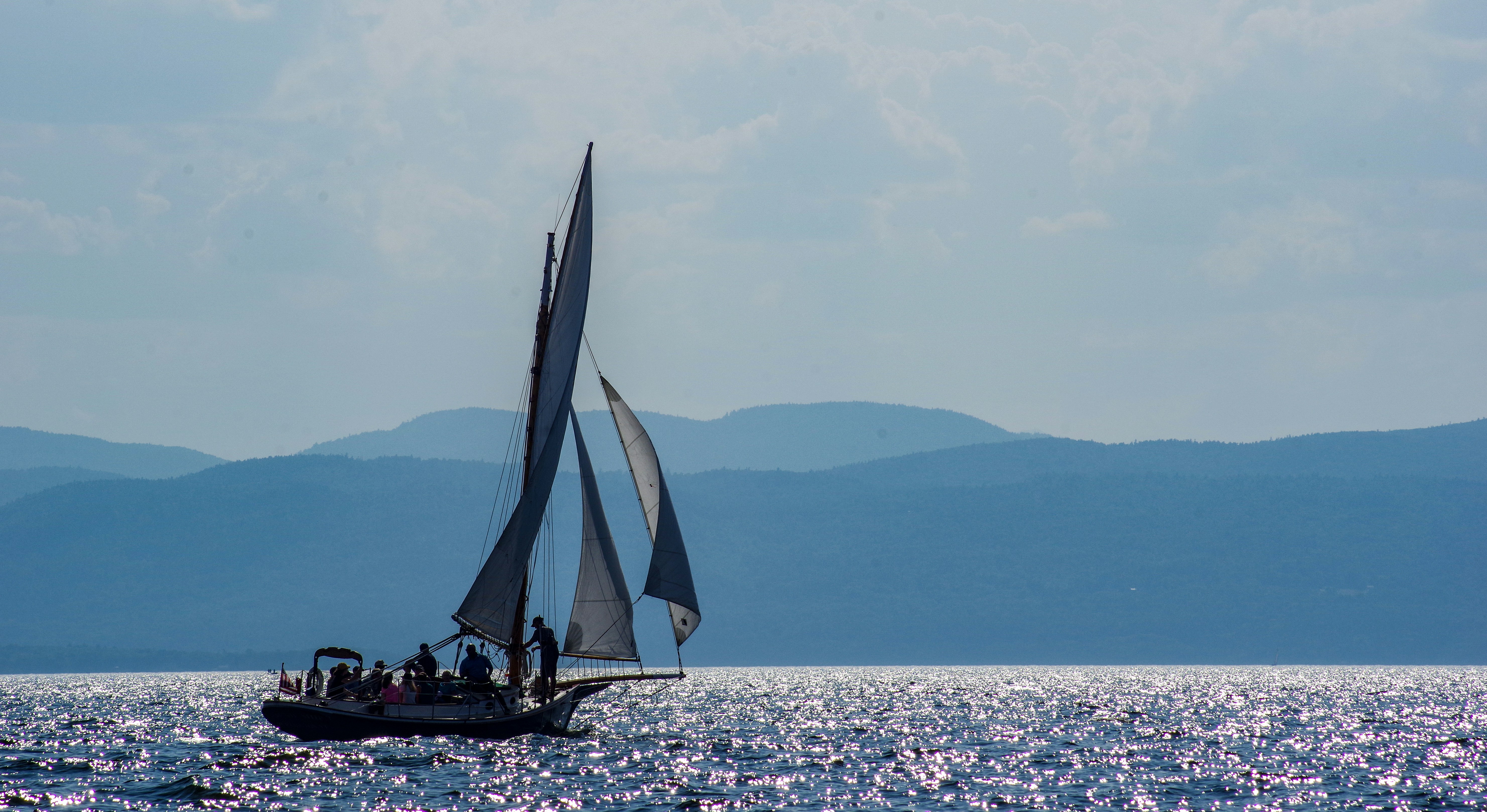 A sailboat with dark sails crossing bright water in front of hazy hills