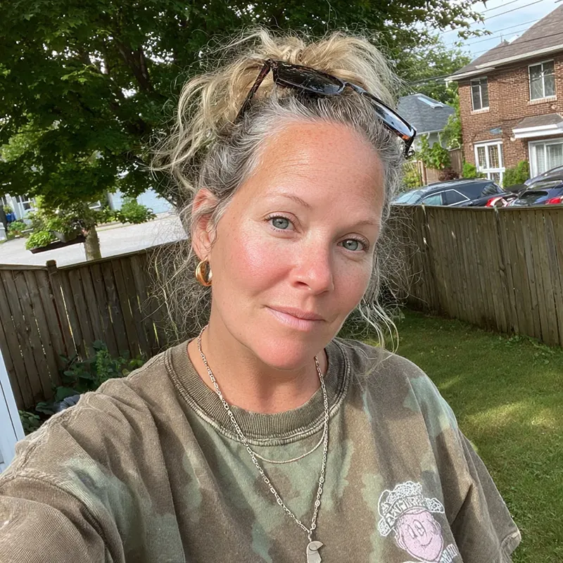 Woman with light hair in a bun, wearing sunglasses and gold hoop earrings, smiles softly in a backyard. Casual vibe with greenery and houses.