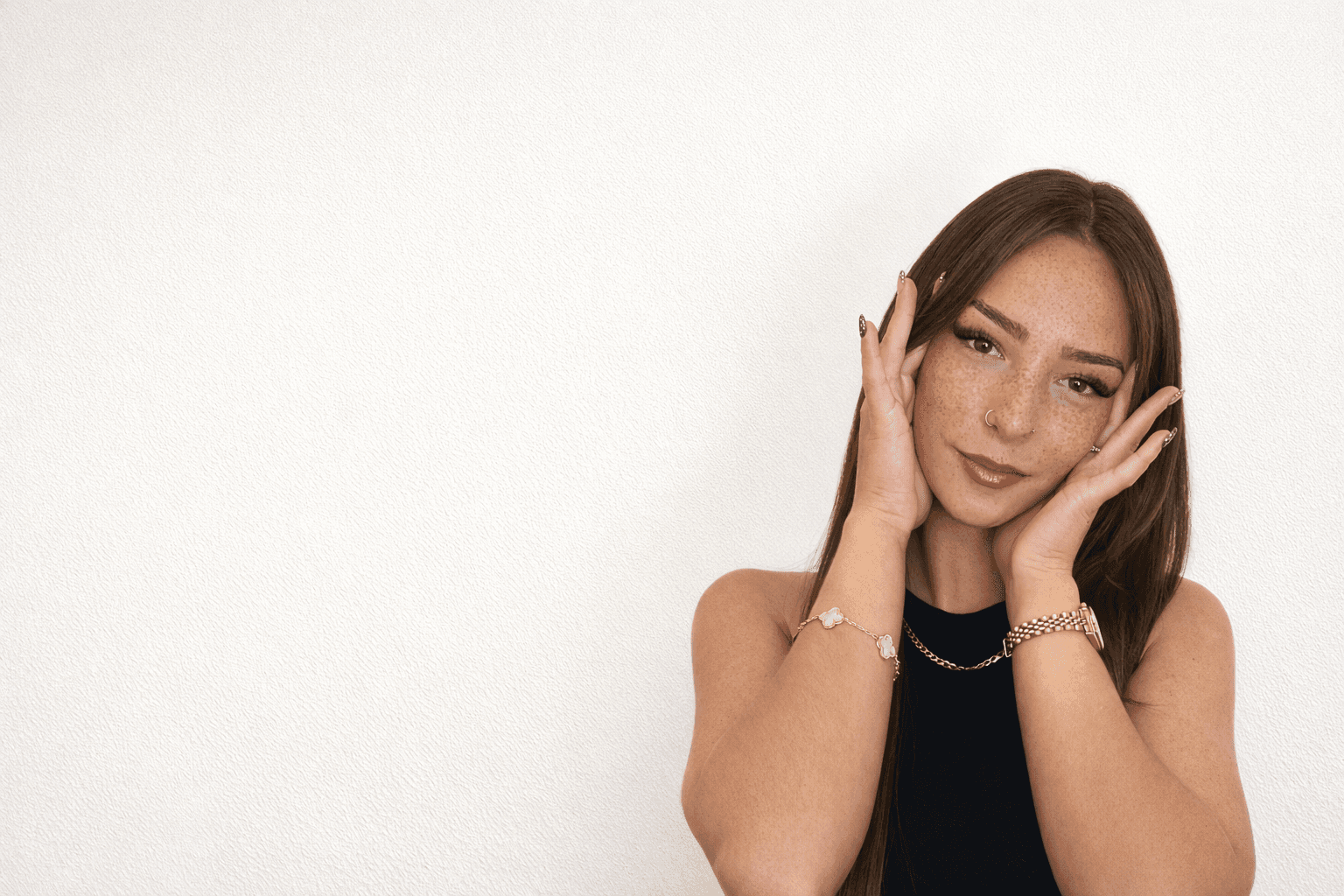 Woman with long brown hair, wearing a black top, covering her ears and looking thoughtfully at the camera against a light background.