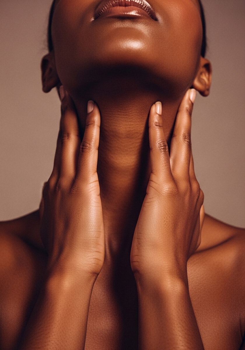 A close-up of a person’s neck and hands, gently touching their throat against a soft, muted background.