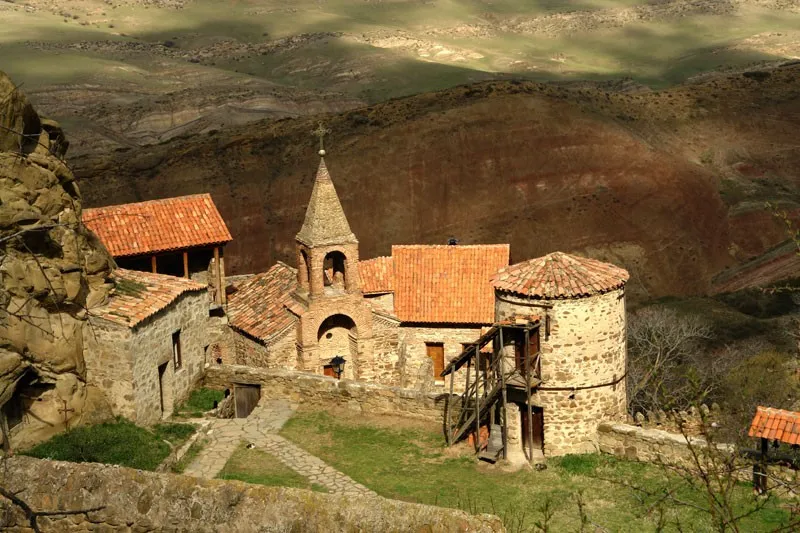 High-angle view of a stone monastery nestled in a hilly landscape. The monastery features terracotta roofs and a bell tower.