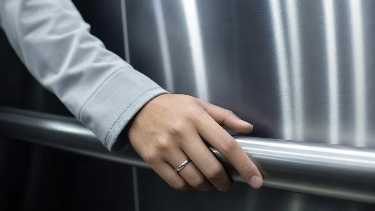 Hand wearing a plain silver dhikr ring resting on an elevator handrail