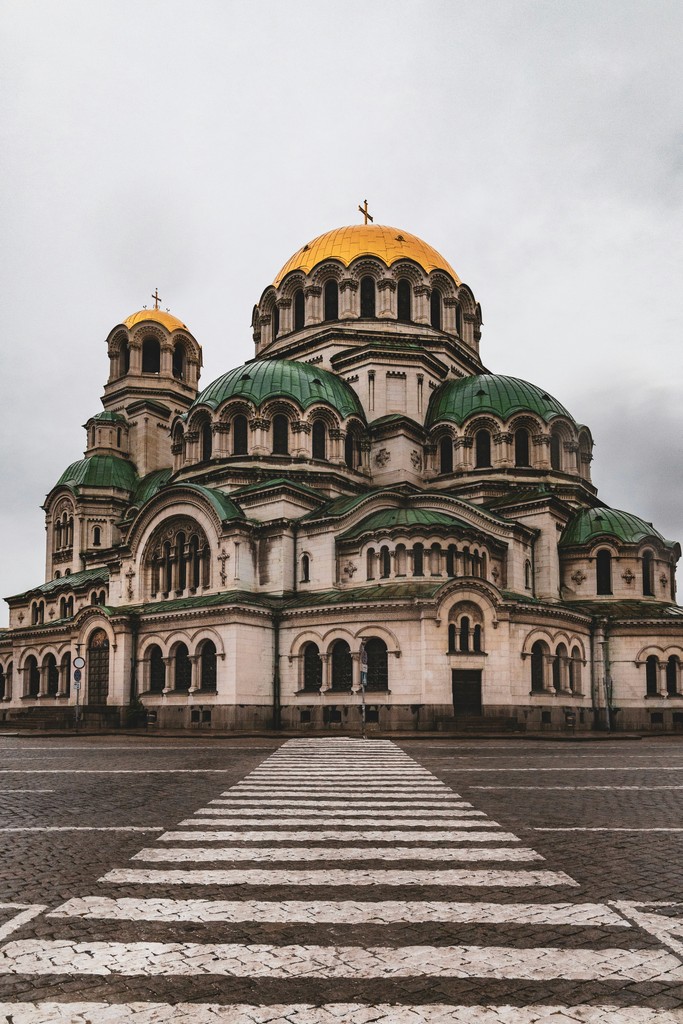 A grand cathedral with a green and gold dome, set against a cloudy sky, with a stone pathway leading up to it.