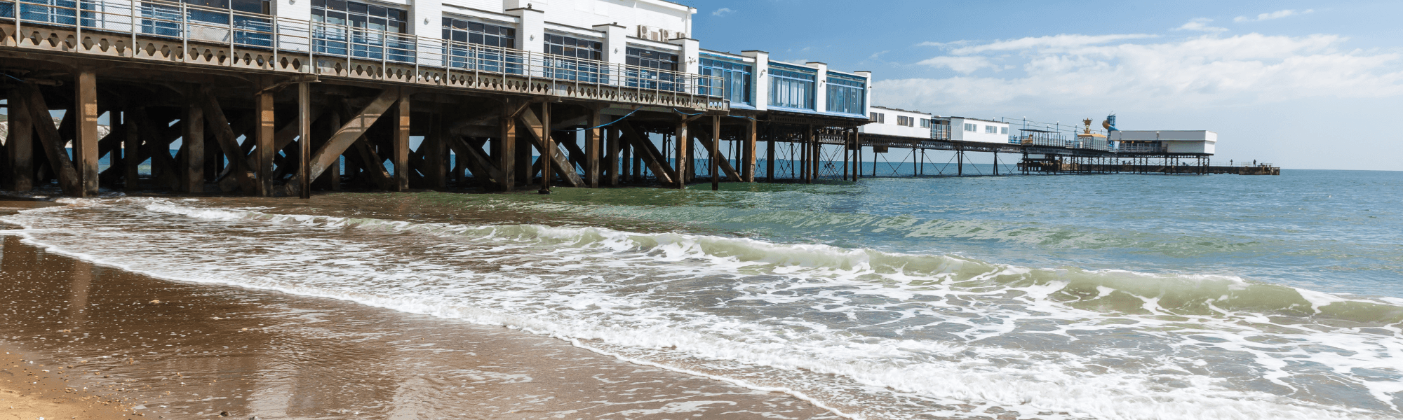A seaside pier extending over the water on a sunny day, with waves breaking on the sandy beach beneath the wooden structure.