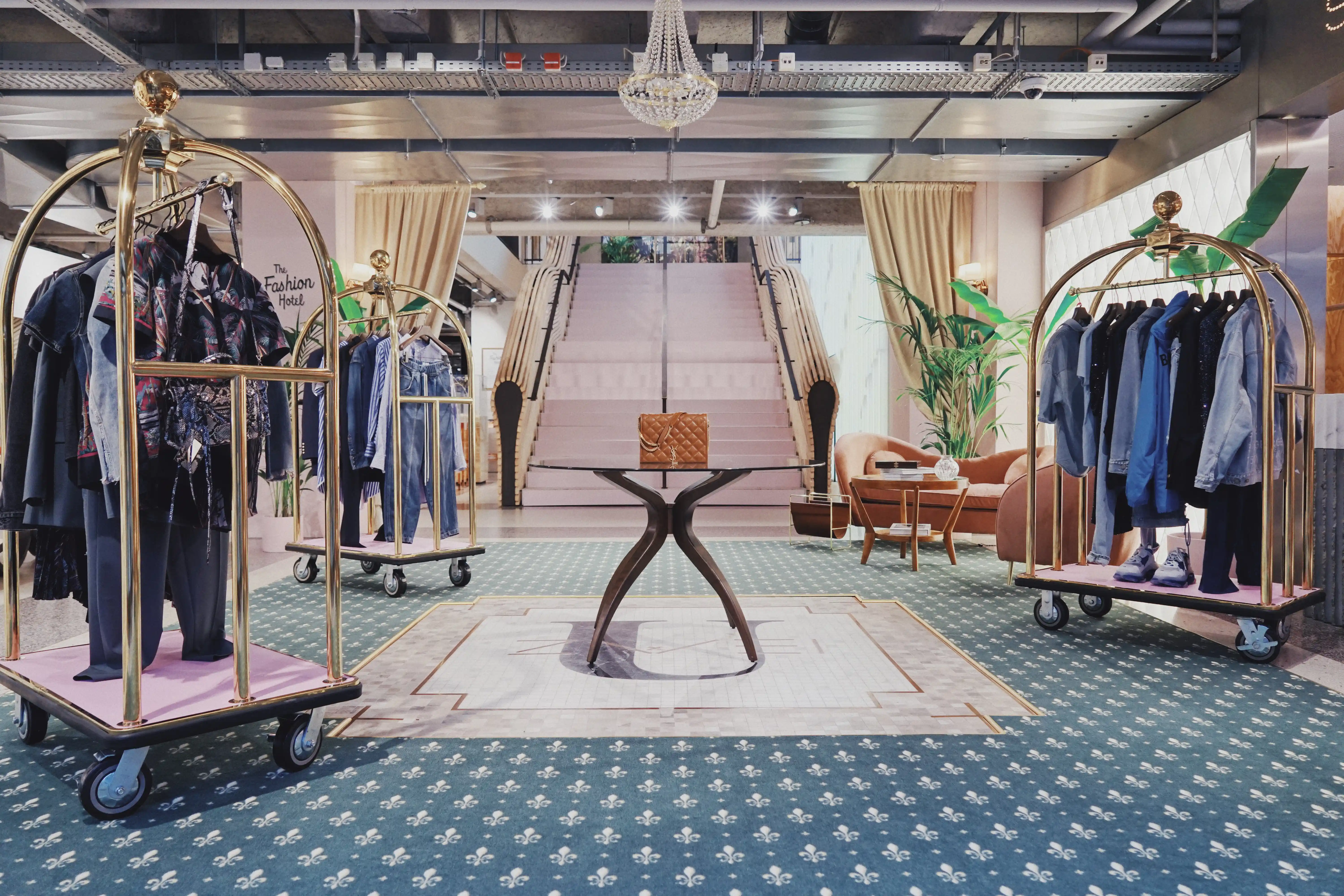 Overview of UZWEI FASHION HOTEL with fleur-de-lis patterned green carpet, a central wooden table with handbag, golden luggage racks with fashion, palms and a chandelier over the staircase.