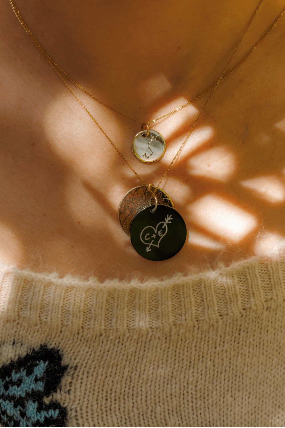 A person wearing three Night Sky pendants. One is a black Onyx Night Sky Love Note of a Heart with an arrow that says "C+E" and it's layered on a Gold Night Sky Star Map. The third pendant is a Scorpius Night Sky Star Sign pendant.