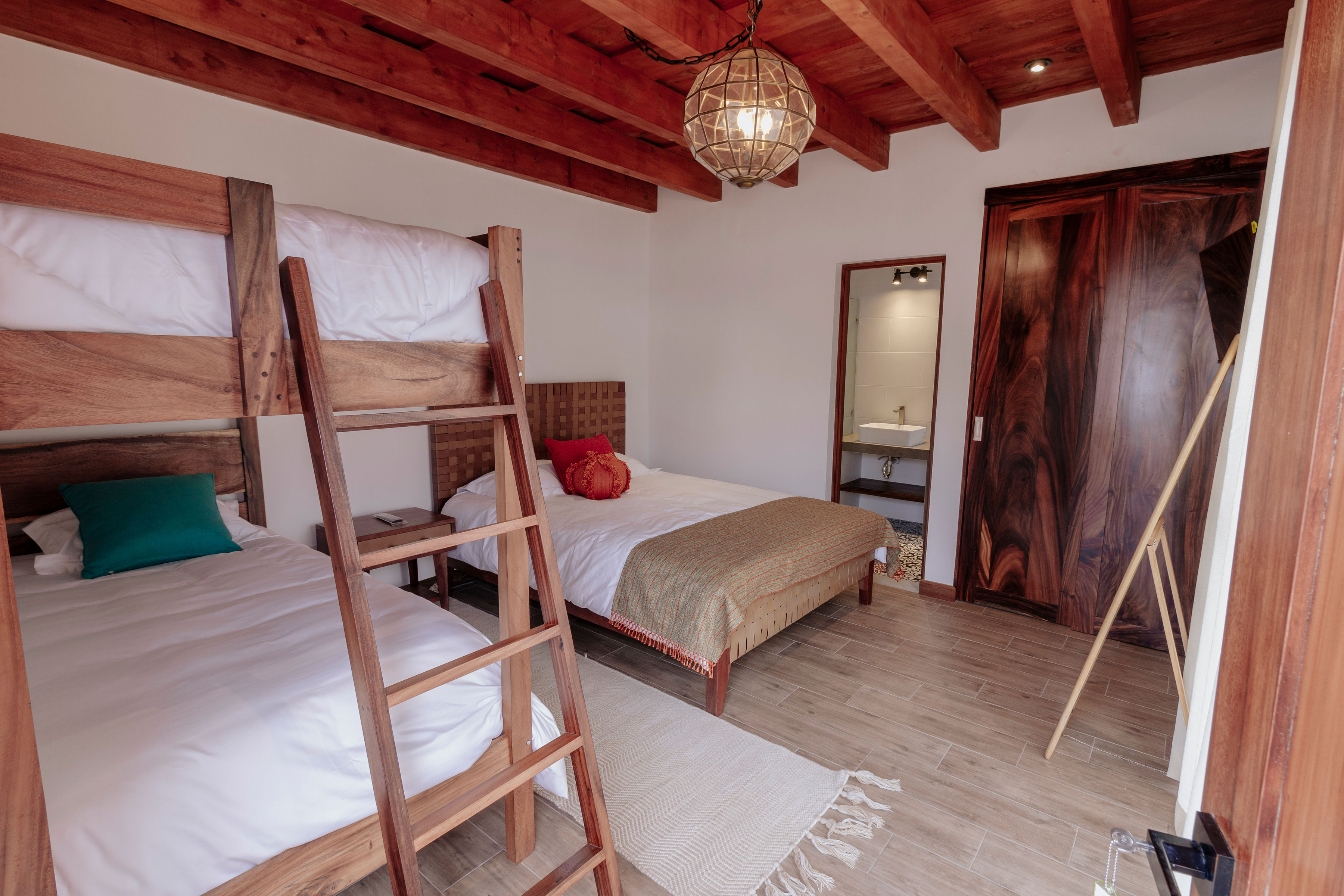 Cozy bedroom with rustic wooden bunk bed and a queen-sized bed, featuring warm wooden ceiling beams, modern decor elements, and a stylish pendant light, creating an inviting and comfortable atmosphere.