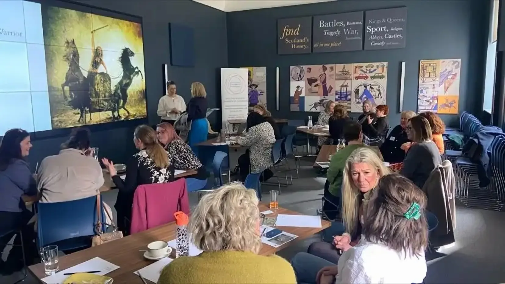 A group of people sits in a well-lit room with dark blue walls and historical posters. They are engaged in discussions at small tables, creating a collaborative and focused atmosphere.