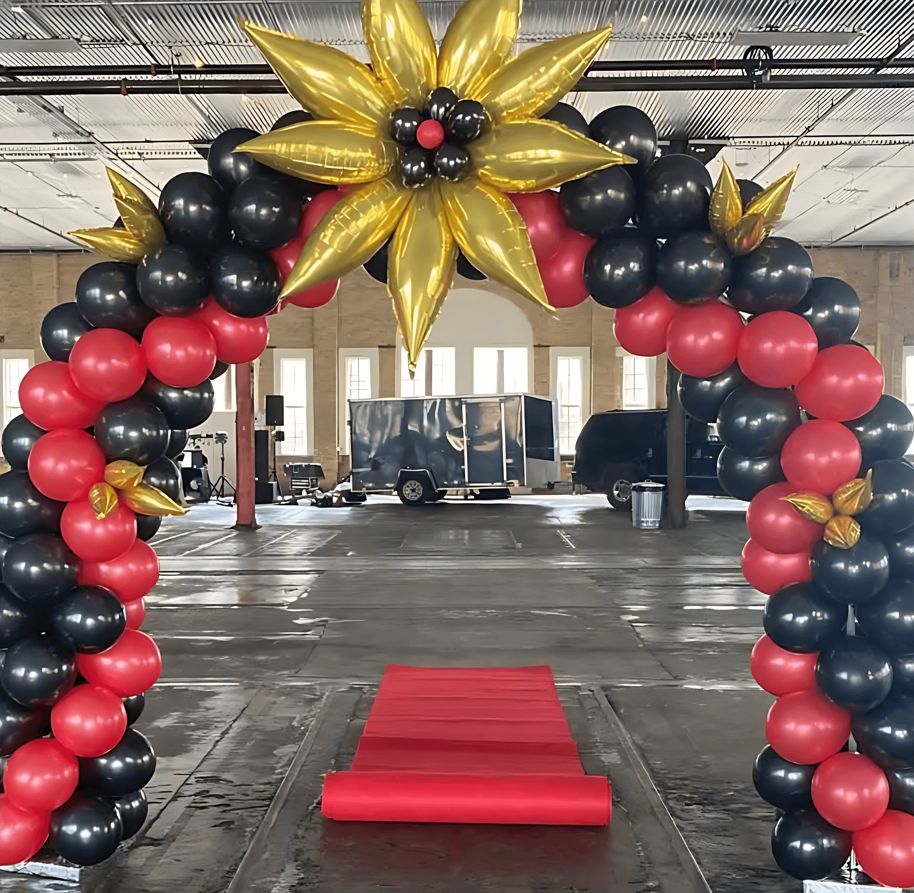 A vibrant balloon arch in black, red, and gold colors frames a red carpet inside a spacious industrial venue, creating a festive and elegant atmosphere.