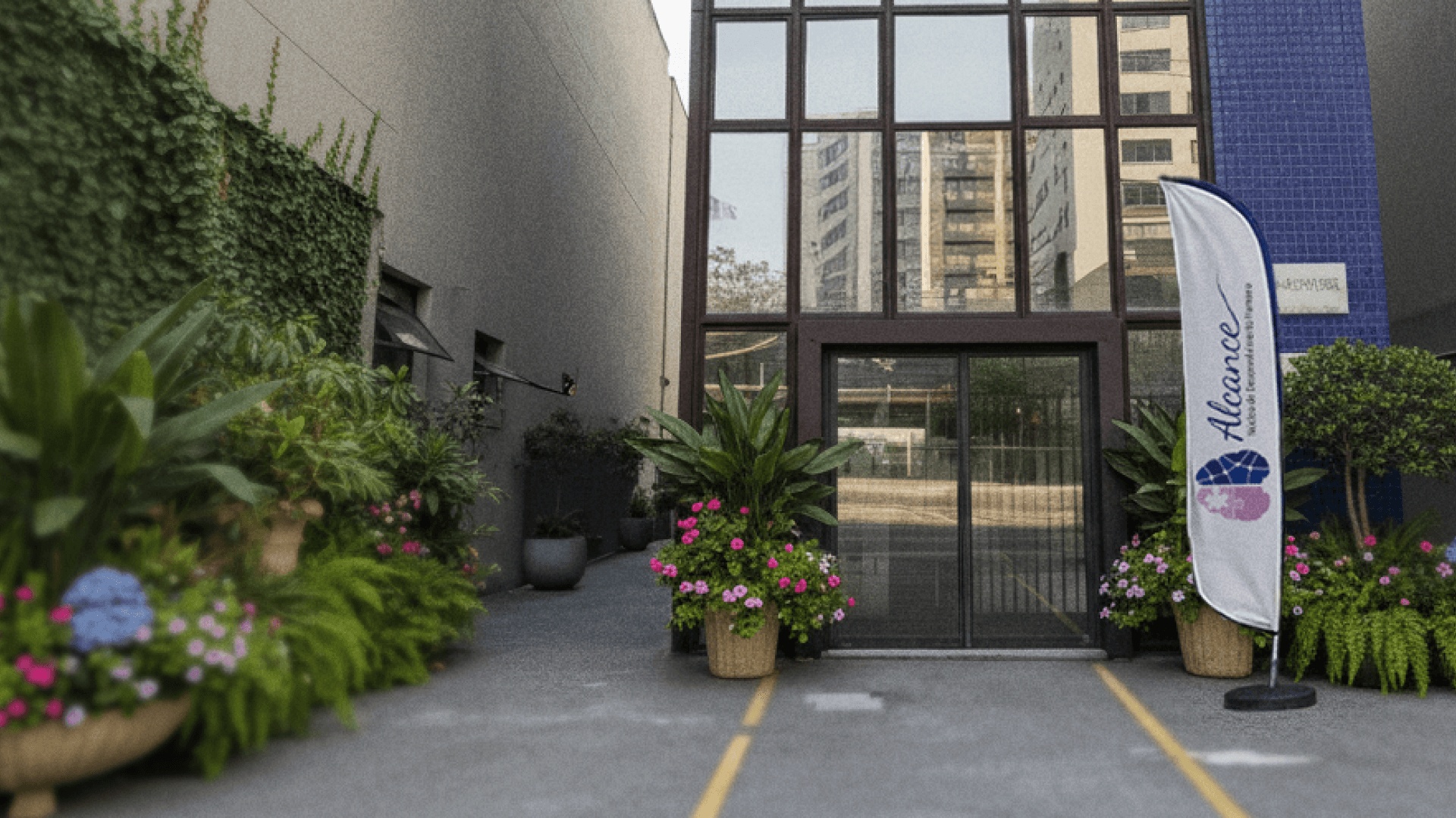 Entrada de um edifício moderno com fachada de vidro e detalhes em pastilhas azuis, cercada por vasos com plantas verdes e flores coloridas. A cena é iluminada por luz natural suave, transmitindo uma sensação de acolhimento, organização e bem-estar em um ambiente urbano e contemporâneo.