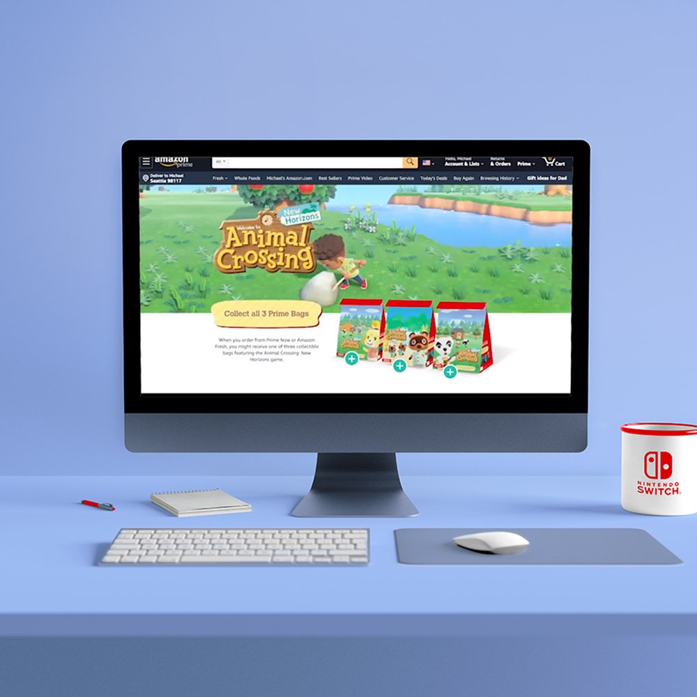 A blue room with a large Mac monitor, white keyboard, and a mouse with a Nintendo coffee mug placed on blue surface. Monitor is showing Animal Crossing New Horizons landing page on Amazon.com