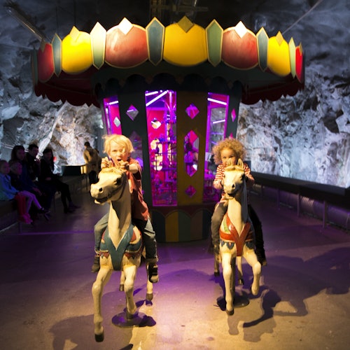 Två barn rider vita karusellhästar på en färgglad, klart upplyst karusell. I bakgrunden syns klippväggar och sittande åskådare.