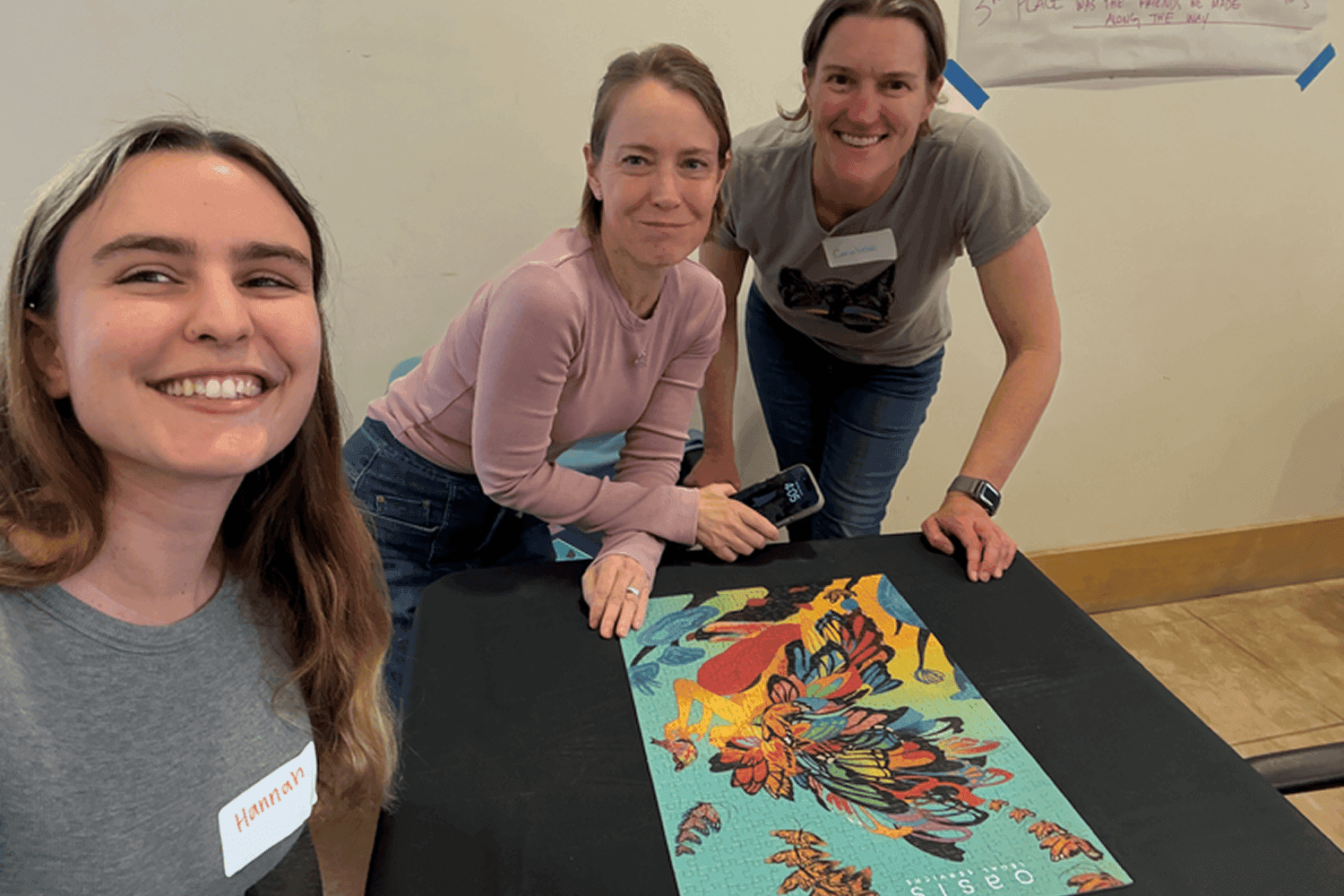 Three individuals are gathered around a table, smiling and completing a colorful, floral-patterned puzzle, with a partially visible poster on the wall in the background.