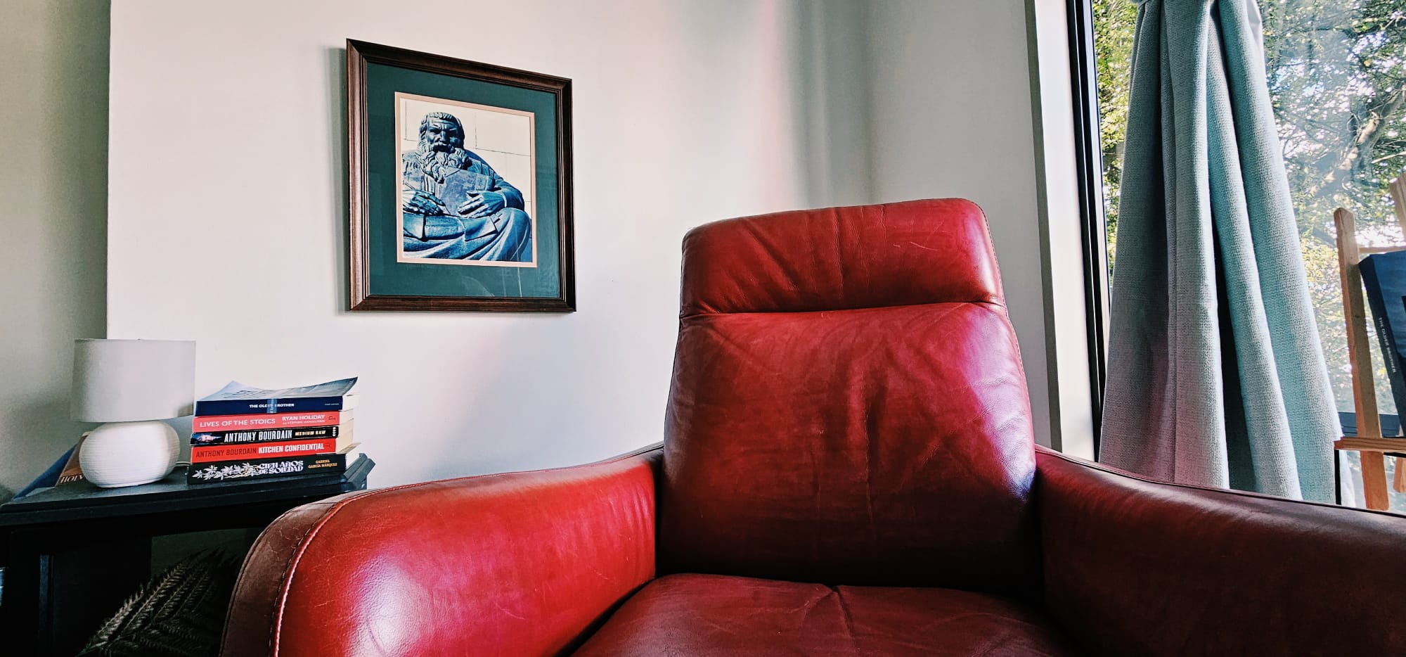 A red reading chair beside a stack of books and a sunlit window in a New Zealand home.