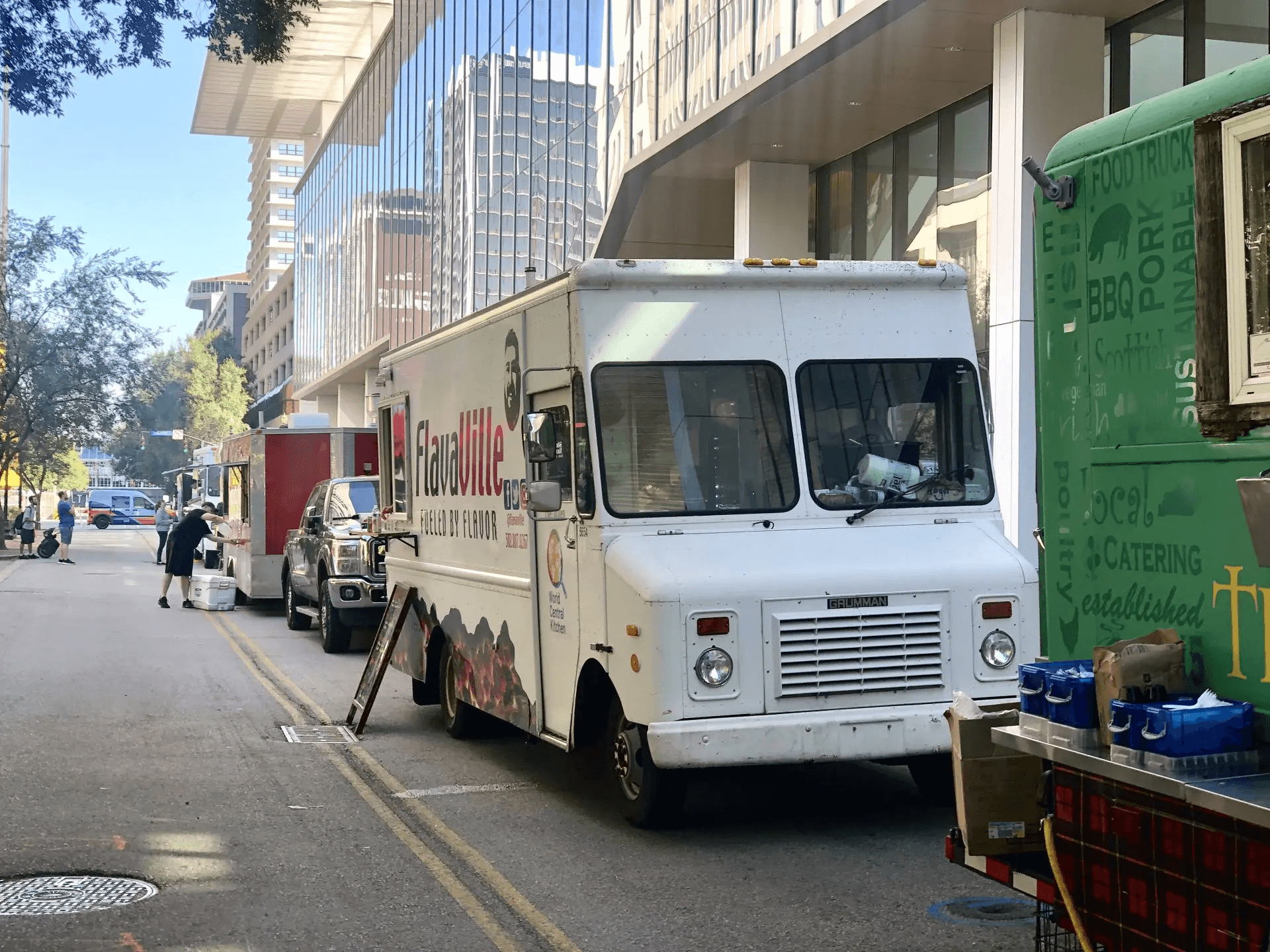 A line of food trucks parked along a city street next to modern glass office buildings, featuring a white truck labeled "FlavaVille Fueled by Flavor."