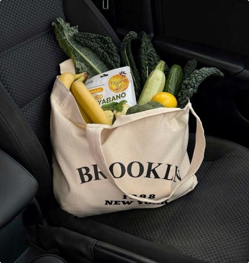 Canvas tote on a car seat filled with fresh produce—bananas, cucumbers, kale, lemons, and a snack pack.
