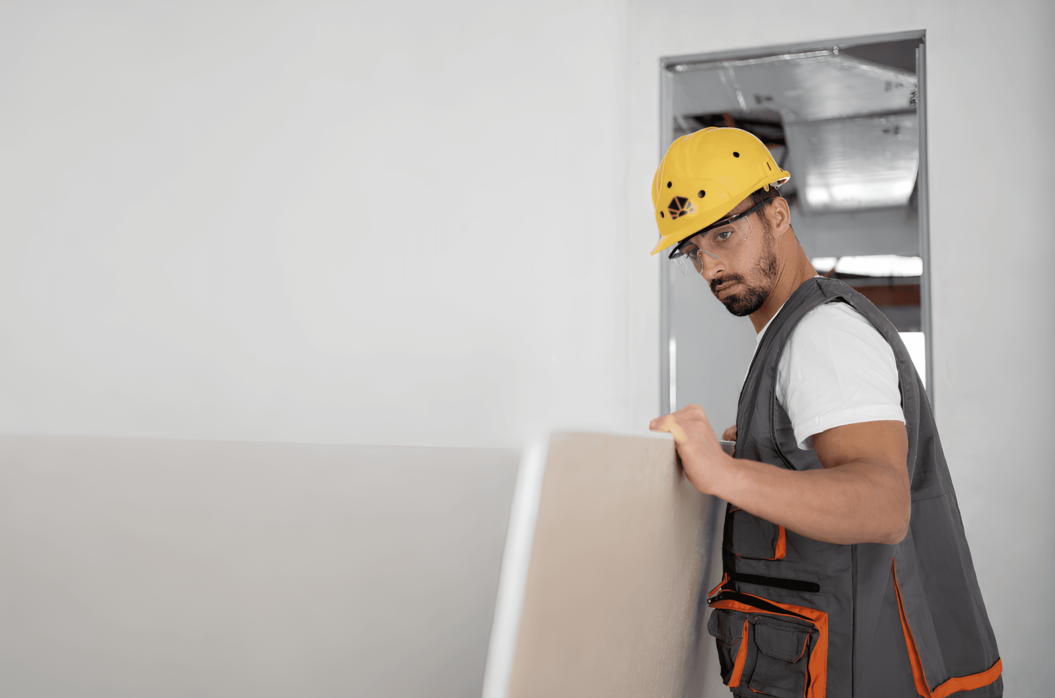 Ein Bauarbeiter mit Schutzhelm und Warnweste trägt Gipskartonplatten in einem Innenbereich.