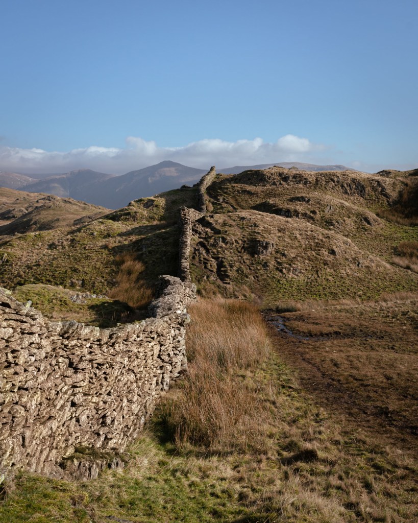 A stone wall leads off into the distance over an undulating terrain. Clear blue sky above with just a few clouds sitting above some distant fells.