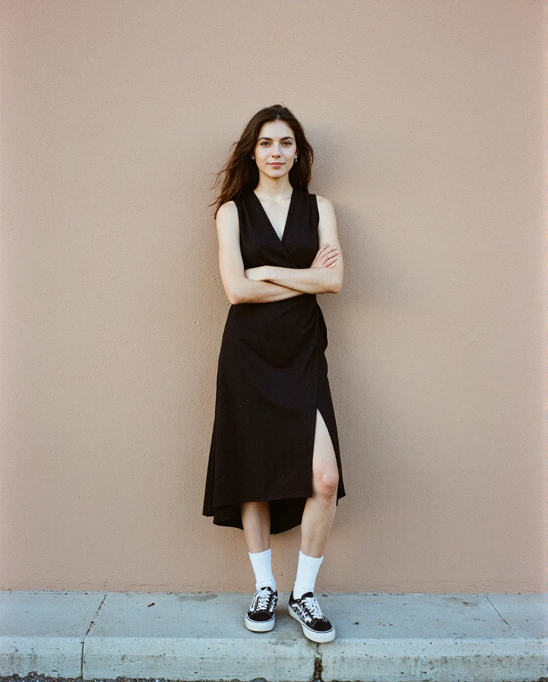 Minimalist fashion portrait featuring a black wrap dress styled with white socks and black sneakers against a neutral beige wall
