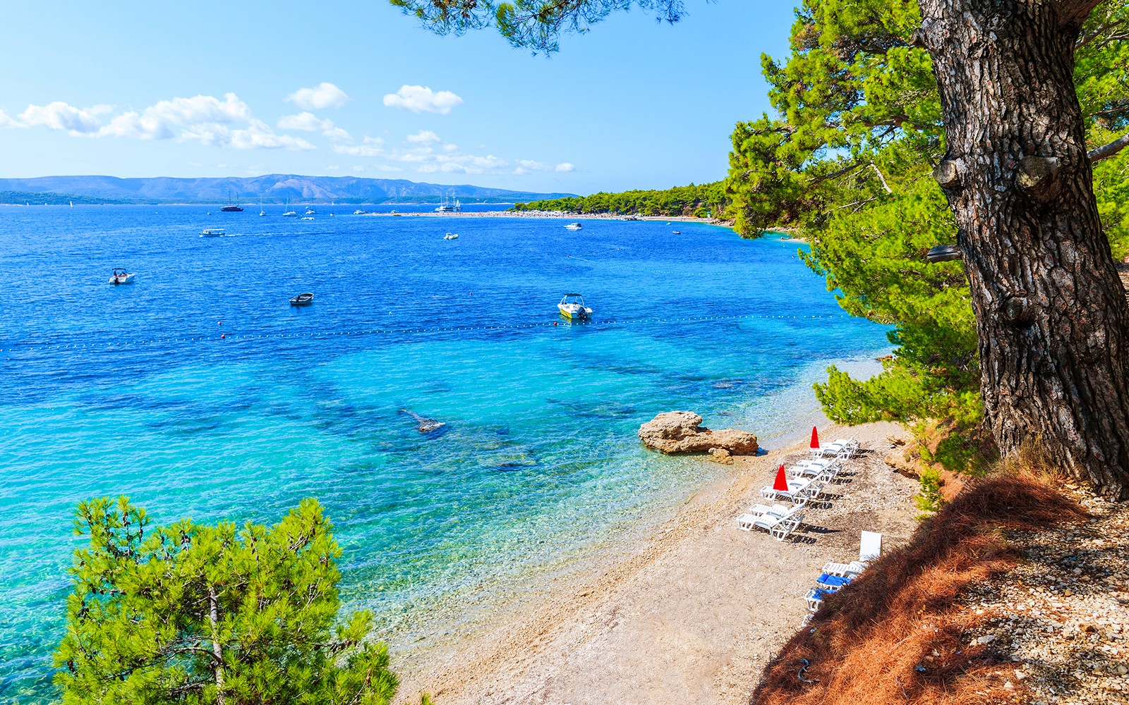 Beach with lounge chairs and umbrellas on Hvar Island, Croatia, overlooking clear blue sea and boats.