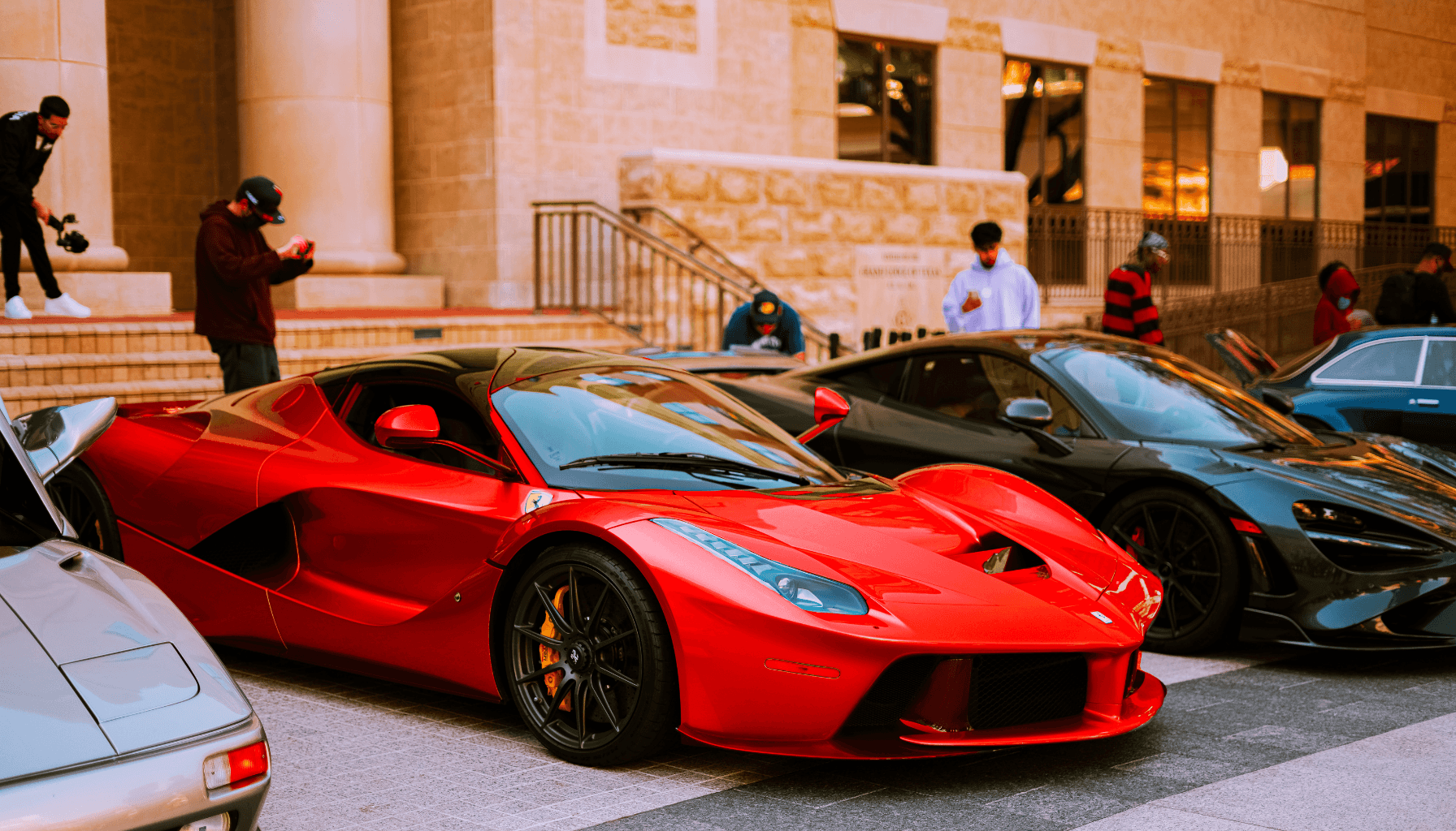 Roter Ferrari und schwarzer Supersportwagen bei einem Autotreffen vor einem modernen Gebäude.
