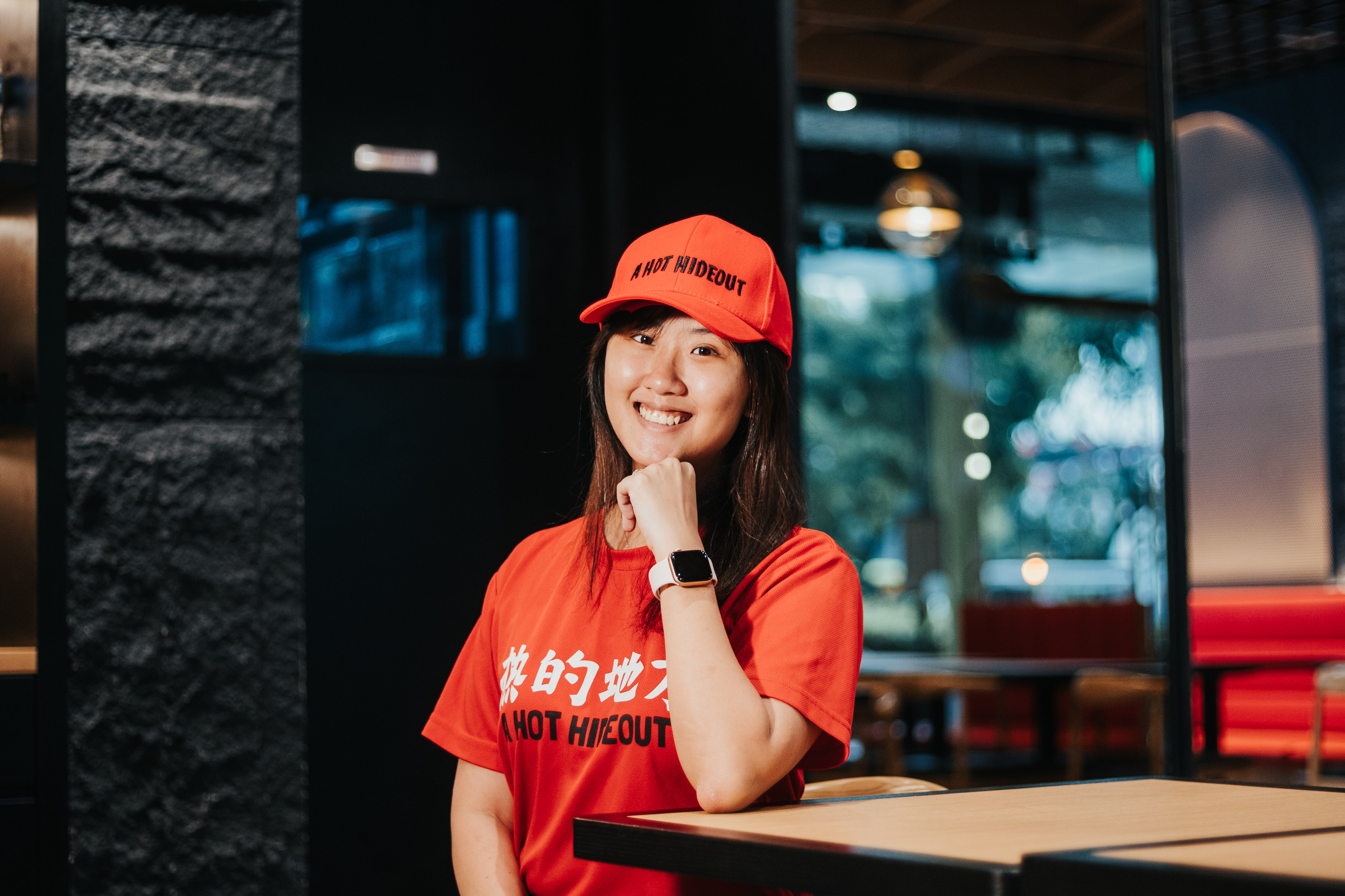 A smiling person in a red cap and shirt stands at a counter in a lively, modern setting. Professional photography.