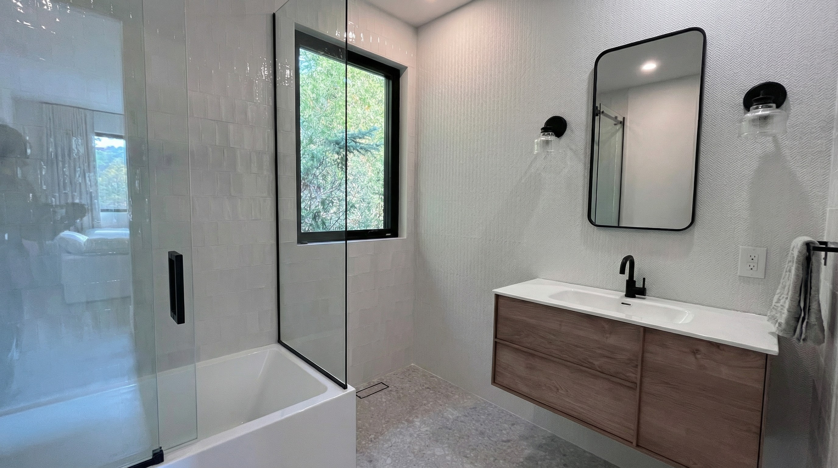 Salle de bain dans les teintes de blanc et de gris. Sur le mur du fond gauche, une céramique blanche lustrée est posée à la verticale, avec une fenêtre au contour noir. On retrouve également un bain douche avec une porte en verre. Sur le mur du fond droit, une mosaïque blanche en forme de chevron. Devant cette mosaïque se trouve une vanité autoportante en bois. Un miroir est installé au mur encadré par deux appliques murales noires, une de chaque côté. Le plancher est recouvert de porcelaine imitant la pierre naturelle grise.