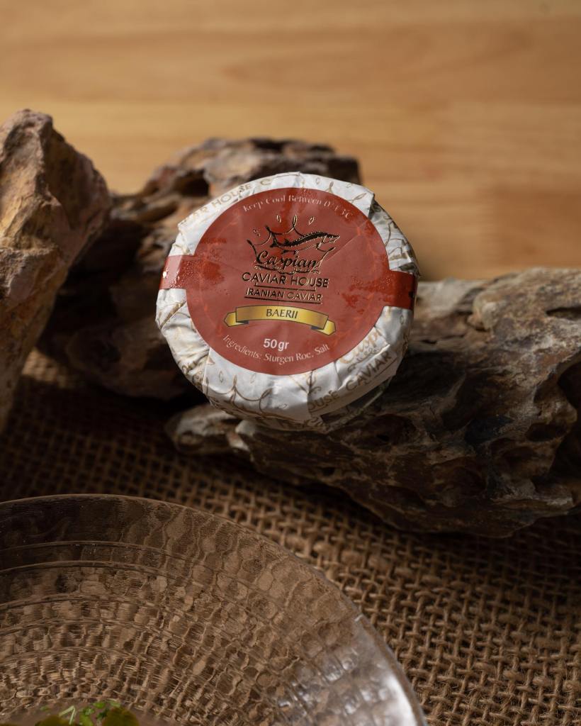 A tin of Caspian Caviar House Ranian caviar labeled as Baerii, placed on rustic rocks, accompanied by a clear glass dish.