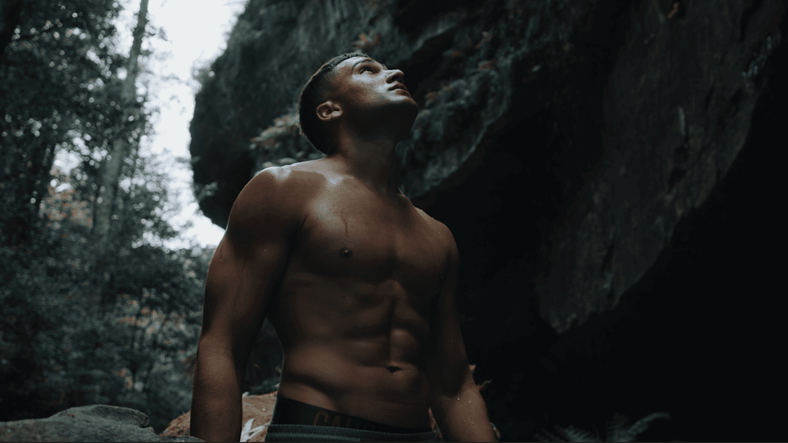 Sweaty muscular man looking up in a rocky, wooded area.