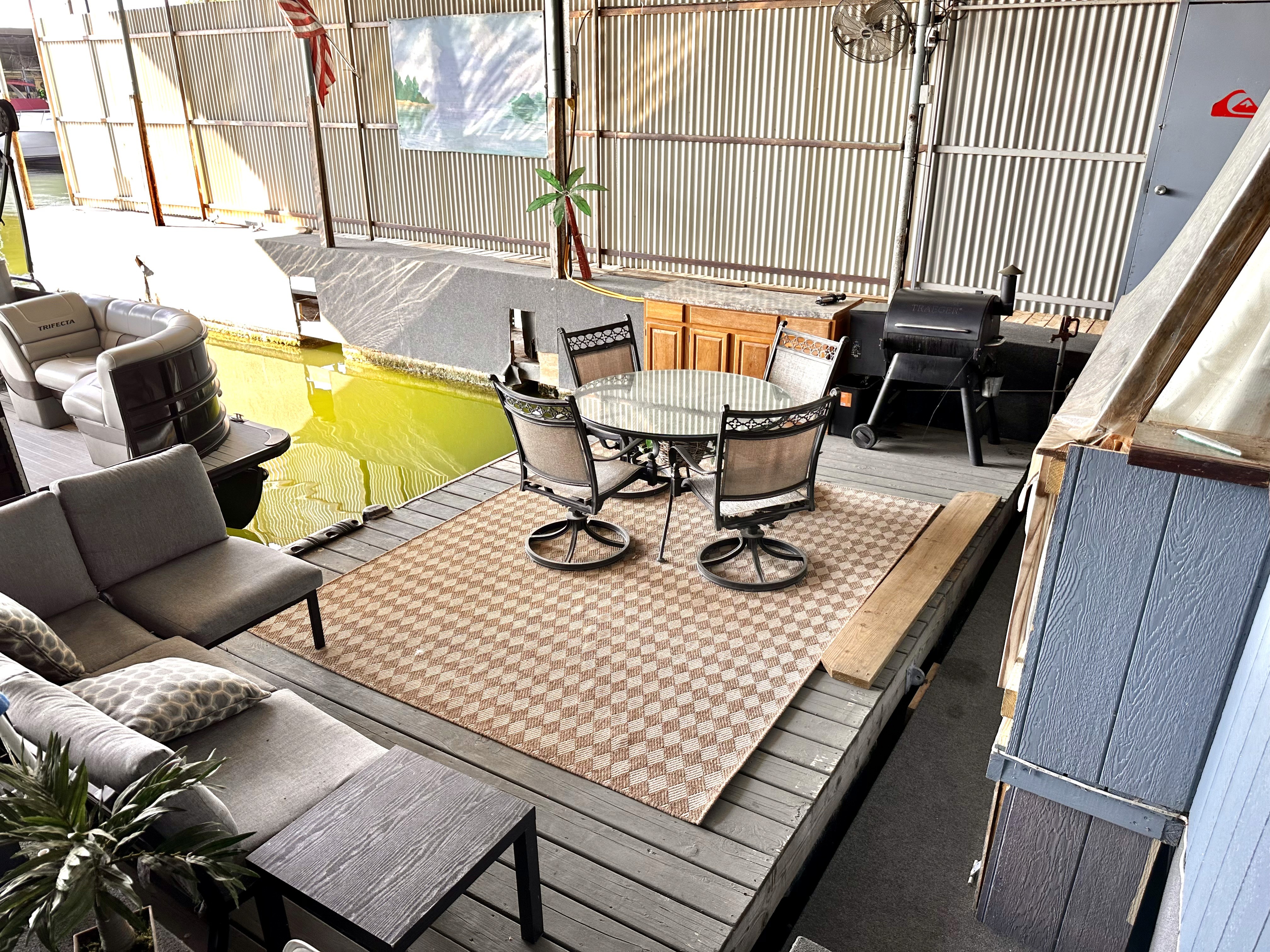 Outdoor seating area with a gray sectional sofa, cushions, and a table set on a wooden deck, featuring a checkered rug, grill, and adjacent yellow waterway against corrugated metal siding.