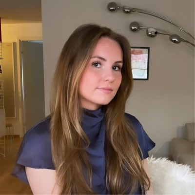 A woman with long hair, wearing sunglasses and smiling on a sunny street with trees in the distance.