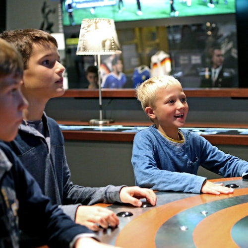 三個男孩正在玩街機遊戲，其中一個在微笑。背景顯示昏暗的燈光和一台顯示足球比賽的電視。