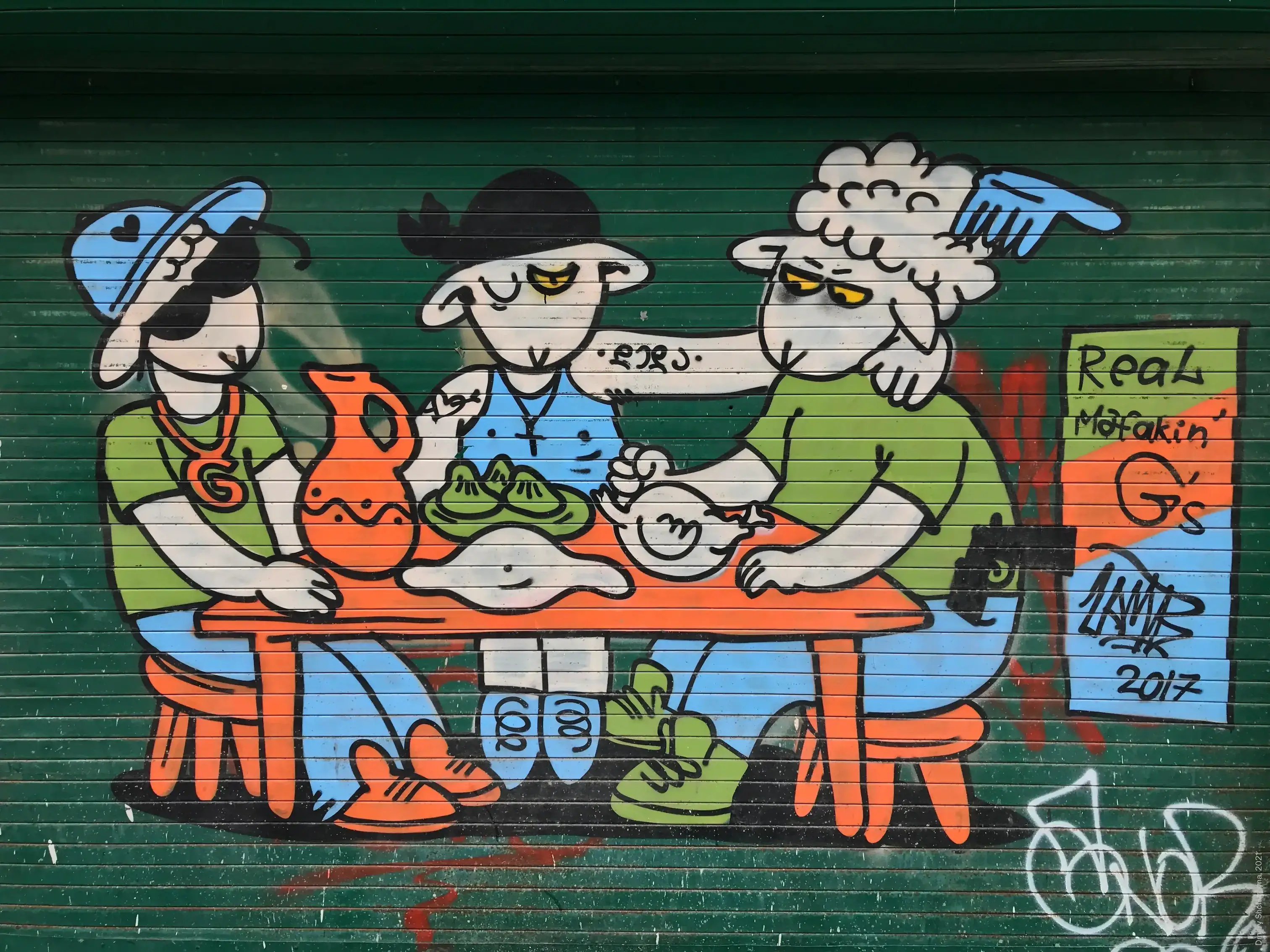 A mural on a green metal roller shutter depicts three cartoon characters with yellow eyes, sitting around an orange table covered in food. They are wearing hats and colorful streetwear.