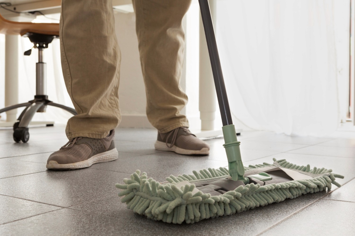 Person cleaning the floor with a green microfiber mop, wearing beige pants and sneakers, showcasing a modern and efficient cleaning method in an office setting.