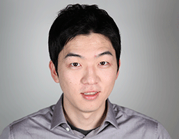 The image shows a man with short, dark hair and dark eyes. He is wearing a light gray collared shirt and appears to be looking directly at the camera. The background is a plain light gray or off-white color. His facial expression is neutral.