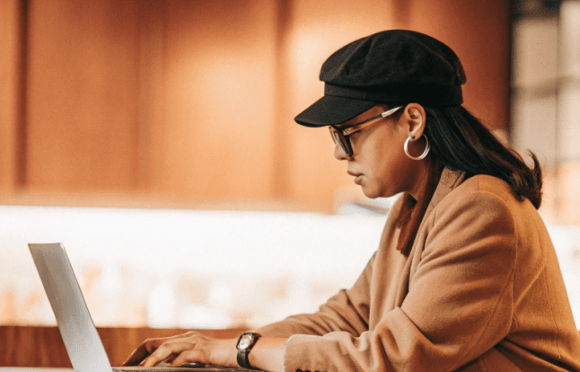 A woman in glasses and a hat works on a laptop, typing. Wearing a blazer and large hoop earrings.