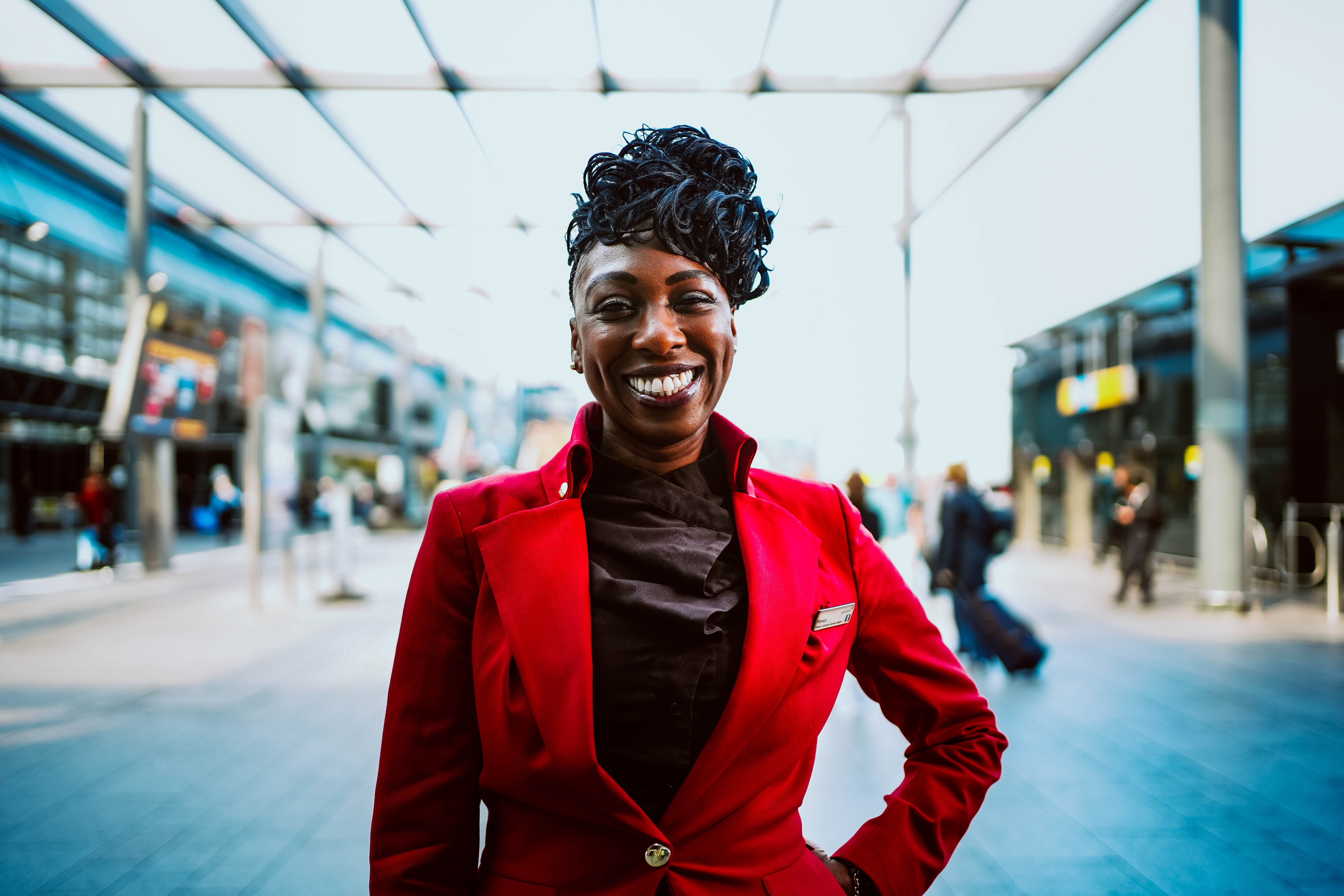 A vibrant portrait of a Black woman in a bright red blazer, smiling warmly at the camera with her hand on her hip. She stands under the modern covered walkway of what appears to be an airport or large station.