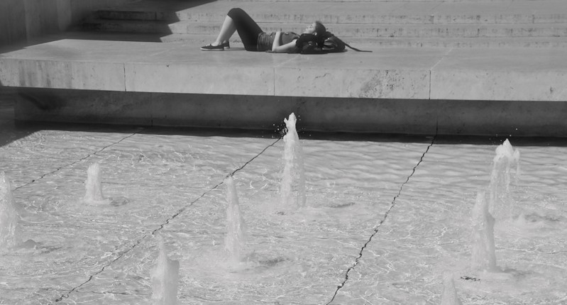 A black and white photo of a fountain spring and a woman which sits next to it and near some stairs.