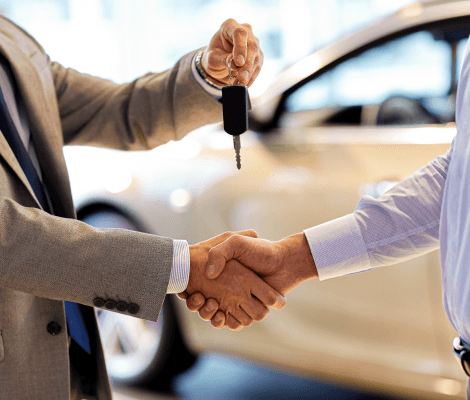 Two individuals are shaking hands in a car dealership setting. One person is handing over a set of car keys while the other is receiving them. A new car is visible in the background, indicating a successful sale or transaction. The atmosphere is professional and celebratory.